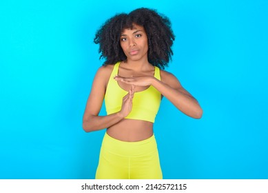 young woman with afro hairstyle in sportswear against blue wall feels tired and bored, making a timeout gesture, needs to stop because of work stress, time concept.