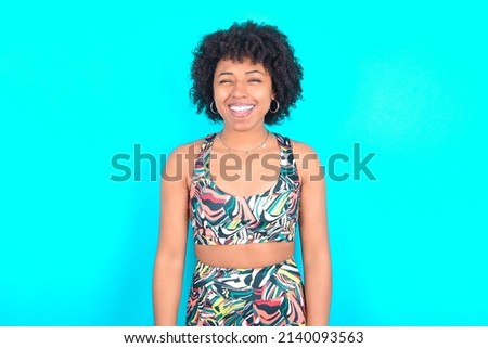 young woman with afro hairstyle in sportswear against blue background blinking eyes with pleasure having happy expression. Facial expressions and people emotions concept.