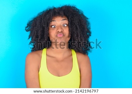 young woman with afro hairstyle in sportswear against blue wall crosses eyes, puts lips, makes grimace with awkward expression has fun alone, plays fool.