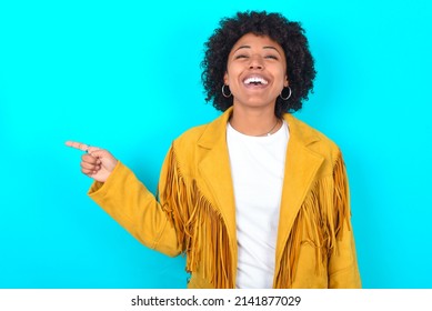 Young Woman With Afro Hairstyle Wearing Yellow Fringe Jacket Over Blue Background Laughs Happily Points Away On Blank Space Demonstrates Shopping Discount Offer, Excited By Good News Or Unexpected Sal