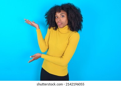 Young woman with afro hairstyle wearing yellow turtleneck over blue background  pointing aside with both hands showing something strange and saying: I don't know what is this. Advertisement concept.