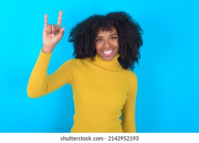 Young Woman With Afro Hairstyle Wearing Yellow Turtleneck Over Blue Background Doing A Rock Gesture And Smiling To The Camera. Ready To Go To Her Favorite Band Concert.