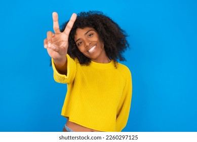 young woman and afro hairstyle wearing yellow sweater against blue background smiling   looking happy  carefree   positive  gesturing victory peace and one hand