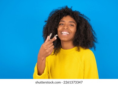 young woman with afro hairstyle wearing yellow sweater against blue background holding an invisible aligner ready to use it. Dental healthcare and confidence concept. - Shutterstock ID 2261373347