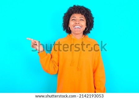 young woman with afro hairstyle wearing orange hoodie against blue background laughs happily points away on blank space demonstrates shopping discount offer, excited by good news or unexpected sale.