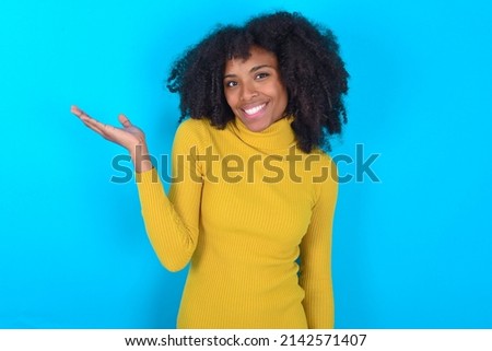 Young woman with afro hairstyle wearing yellow turtleneck over blue background smiling cheerful presenting and pointing with palm of hand looking at the camera.
