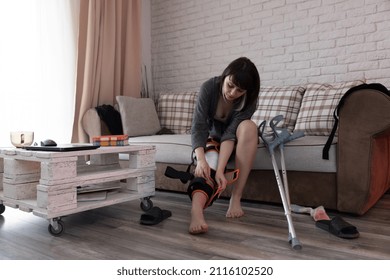 Young Woman After An ACL Surgery, Sitting On The Couch Adjusting Her Knee Orthosis.