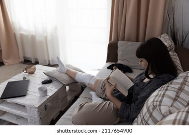 Young Woman After An ACL Surgery, Sitting On The Couch And Reading A Book.