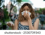 Young woman with allergies in a park, blowing her nose due to poplar fluff, looking determined despite feeling unwell