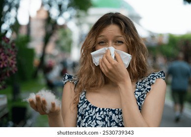 Young woman with allergies in a park, blowing her nose due to poplar fluff, looking determined despite feeling unwell - Powered by Shutterstock