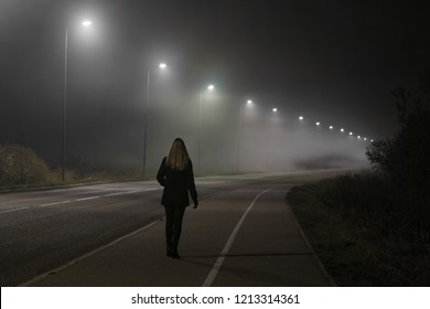 Young woman alone slowly walking under white street lights in night. Dark time. Peaceful atmosphere in mist. Foggy air. Back view.