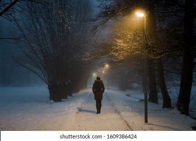 Young woman alone slowly walking on sidewalk under yellow street lights in mist. Foggy air. Peaceful atmosphere in snowy night. Back view.