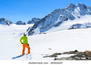 Young Woman Alpine Skier In Orange Pants And Green Jacket Stands In Fresh Snow On Pitztal Glacier In Austrian Alps