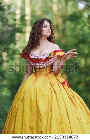 A young woman in an ancient medieval yellow orange dress walks in a green park on a summer day