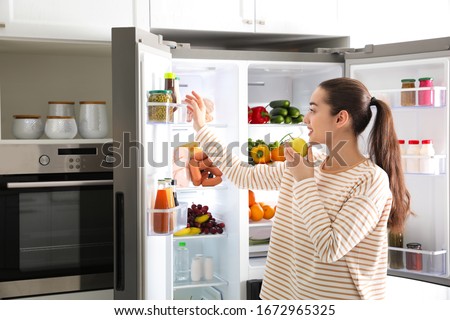 Young woman with apple near open refrigerator in kitchen