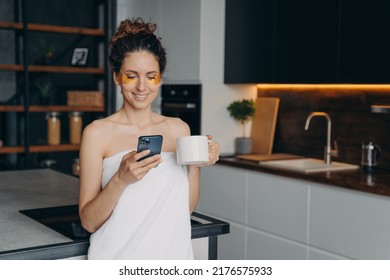 Young woman applies anti wrinkle eye patches and relaxing at home texting on phone. Happy hispanic girl wrapped in towel after spa procedures. Body care, beauty and wellness. Modern interior. - Powered by Shutterstock