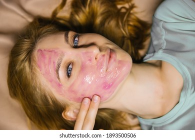 A Young Woman Applies A Pink Cosmetic Mask Or Gel To Her Face. Facial Skin Care. The Girl Smiles And Looks At The Camera.