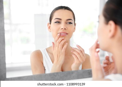 Young Woman Applying Balm On Her Lips Near Mirror Indoors