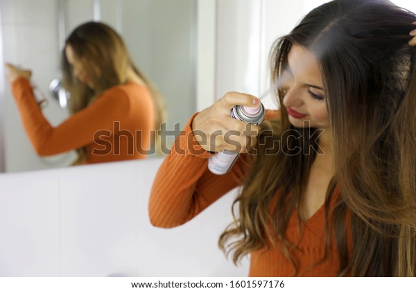 Young woman applying dry shampoo on her hair. Fast\
and easy way to keep hair clean with dry shampoo. Focus on the hand\
with spray.