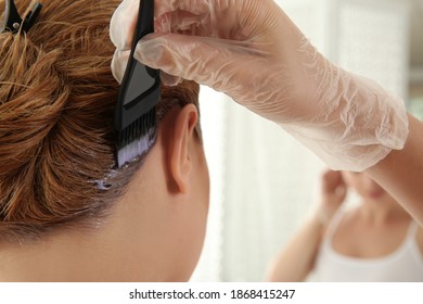 Young woman applying dye on hairs near mirror indoors, closeup