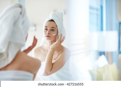 Young Woman Applying Foundation Or Moisturizer On Her Face In Front Of Mirror