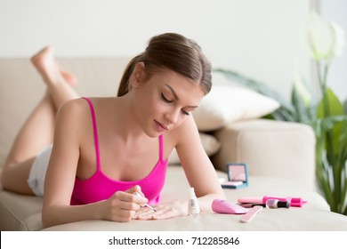 Young woman applying lacquer by brush from bottle, polishing painting fingernails with colorless protective enamel while lying on sofa, doing french manicure at home, perfect healthy nails care - Powered by Shutterstock