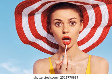 Young woman applying lipstick while wearing a large striped sun hat against a bright blue sky - Powered by Shutterstock