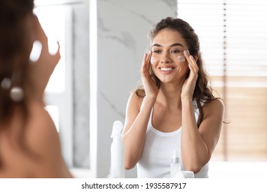 Young Woman Applying Moisturizing Facial Cream Looking In Mirror Indoor - Powered by Shutterstock