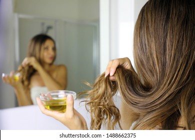 Young Woman Applying Olive Oil Mask To Hair Tips In Front Of A Mirror. Haircare Concept. Focus On Hair.