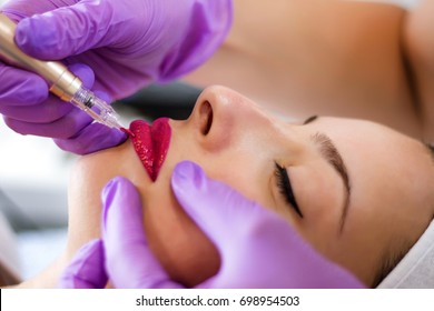 Young woman applying permanent make up on lips in beautician salon with tattoo tool. Selective focus close up - Shutterstock ID 698954503