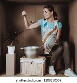 Young Woman In Apron Cooking In Cardboard Box. Life In Little Cardboard Box. Uncomfortable Life Concept. Personal Spase Concepts. Sitting Girl. Girl In Apron. Cooking Food. Smiling Woman.