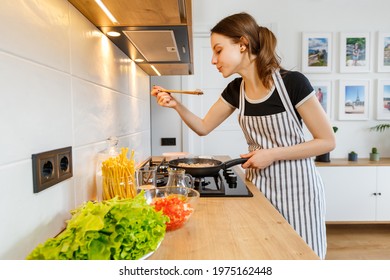 Young Woman In Apron Cooking Healthy Food At Modern Home Kitchen. Preparing Meal With Frying Pan On Gas Stove. Concept Of Domestic Lifestyle, Happy Housewife Leisure And Culinary Hobby.