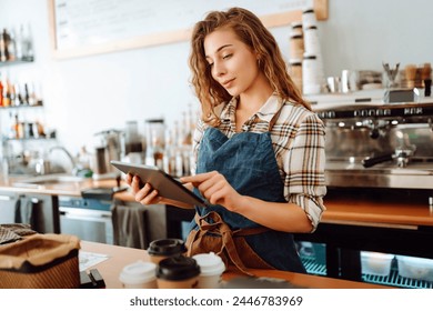 A young woman in an apron with a digital tablet at the bar counter of a coffee shop takes an order. Small business. Food and drinks. - Powered by Shutterstock