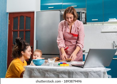 A young woman in an apron makes cookies. There is a laptop on the table. A baby is sitting on a chair next to me, in the arms of a young woman. The concept of home cooking and modern technologies.