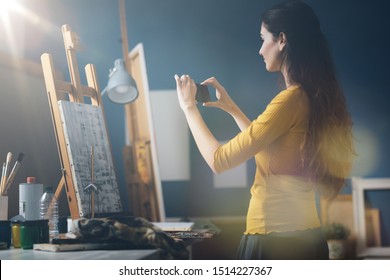 Young woman in the art studio taking a picture of her painting using her smartphone, art and technology concept - Powered by Shutterstock