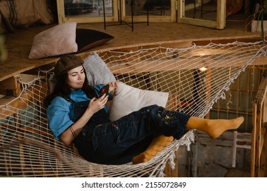 Young woman artist using cellphone while resting in hammock at her studio indoors - Powered by Shutterstock