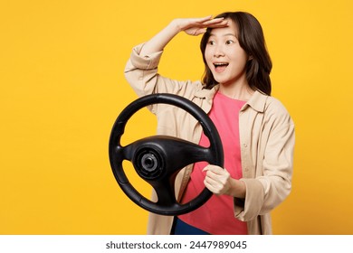 Young woman of Asian ethnicity wear pink t-shirt beige shirt pastel casual clothes hold steering wheel driving car look far away isolated on plain yellow background studio portrait. Lifestyle portrait - Powered by Shutterstock