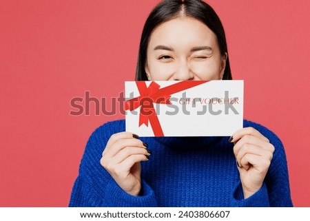 Young woman of Asian ethnicity wear blue sweater casual clothes hold cover mouth with gift certificate coupon voucher card for store isolated on plain pastel light pink background. Lifestyle concept