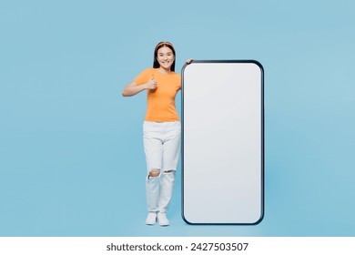 Young woman of Asian ethnicity wears orange t-shirt casual clothes big huge blank screen mobile cell phone smartphone with workspace copy space mockup show thumb up isolated on plain blue background