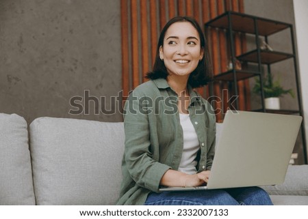 Young IT woman of Asian ethnicity wears casual clothes work hold use laptop pc computer look aside sit on grey sofa couch stay at home hotel flat rest relax spend free spare time in living room indoor