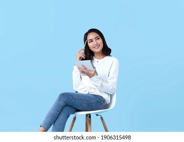 Young woman Asian happy and interested in thinking.While her holding notebook sitting on white chair isolate on bright blue background. - Shutterstock ID 1906315498