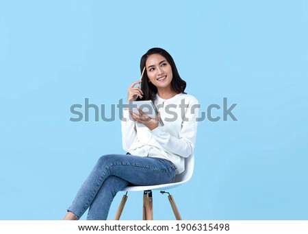 Young woman Asian happy and interested in thinking.While her holding notebook sitting on white chair isolate on bright blue background.