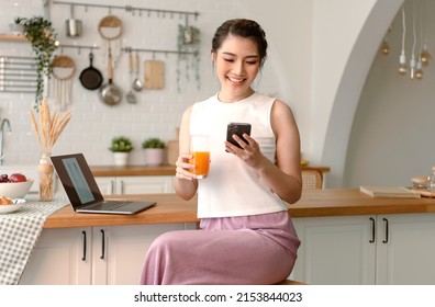 Young woman Asian happy rest drinking orange juice and using mobile phone in kitchen room. - Powered by Shutterstock