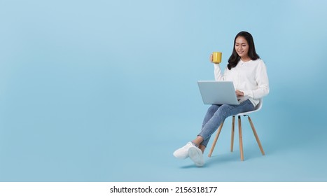 Young woman asian happy smiling in casual white cardigan with denim jeans.While her using computer laptop sitting on white chair and drinking coffee isolate on bright blue background. - Powered by Shutterstock