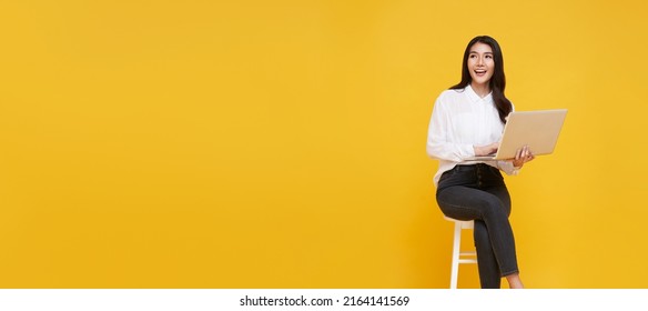 Young woman asian happy smiling. While her using laptop sitting on white chair and looking isolate on copy space yellow background. - Shutterstock ID 2164141569