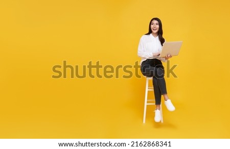 Young woman asian happy smiling. While her using laptop sitting on white chair and looking isolate on copy space yellow background.