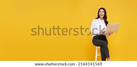 Young woman asian happy smiling. While her using laptop sitting on white chair and looking isolate on copy space yellow background.
