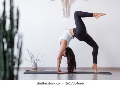 Young woman asian yoga lover doing standing in Bridge exercise, One legged Wheel pose on floor in cozy white room. Working out, wearing sportswear, black pants, bra, indoor full length, home interior