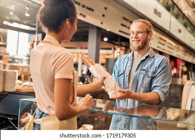 Young woman assistant wearing apron giving packed wheat baguette to smiling customer at bakery shop small business - Powered by Shutterstock