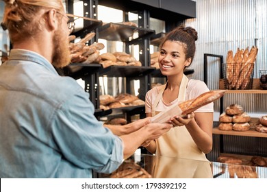 Young woman assistant wearing apron giving packed wheat baguette to customer at bakery shop small business smiling happy - Powered by Shutterstock